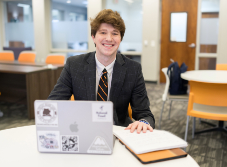 male student dressed in business professional with a laptop and notebook on table in front of him